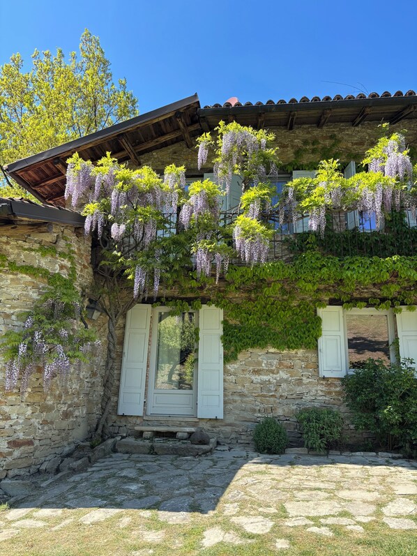 Ein Gebäude aus Stein mit einem Holzdach, weißen Fensterläden und hängenden, violetten Blumen.