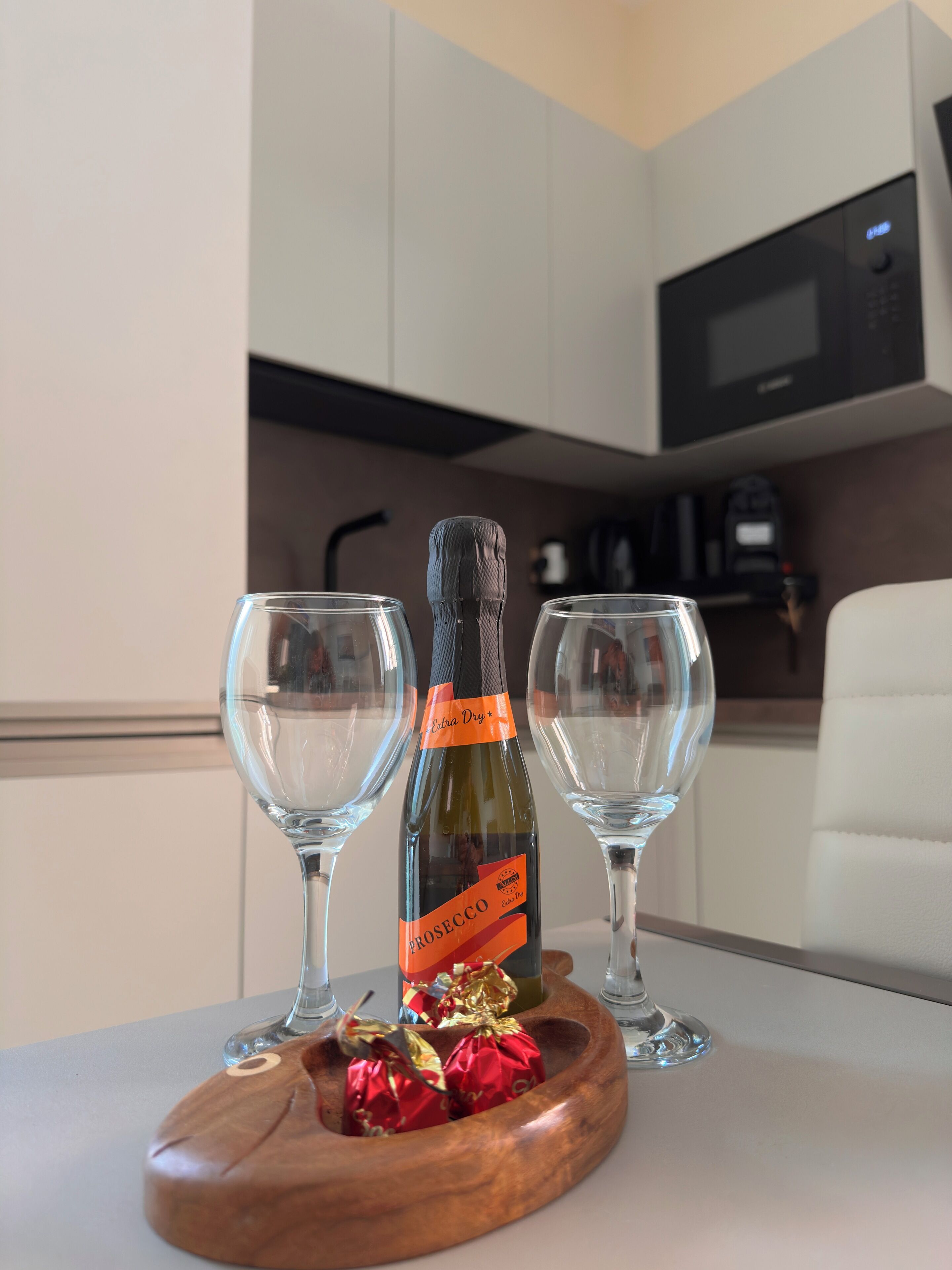 A bottle of Prosecco wine on a table with two empty wine glasses and a wooden coaster with wrapped chocolates.