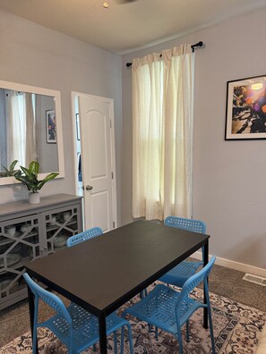 Un'area pranzo con un tavolo in legno scuro, sedie blu, una credenza con stoviglie, uno specchio e una finestra con tende.
