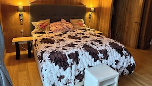 Quarto com painéis de madeira, uma cama grande, duas luminárias de cabeceira e um banquinho branco pequeno.