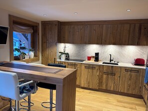 A modern kitchen with wooden cabinets, a dining area with a bar stool, and a window with plants. - MONT ARBOIS MEGEVE, apartment on the slopes (Megève)