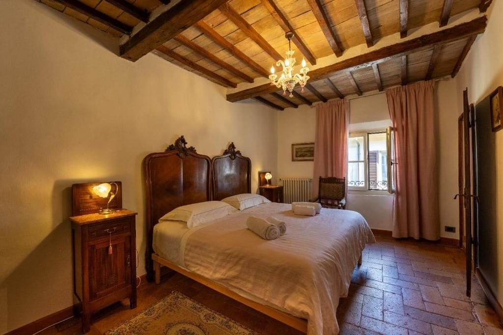 Ein Schlafzimmer mit Holzboden, einem Bett mit weißen Leinen, einem Nachttisch mit Lampe und einem Fenster mit Vorhängen.