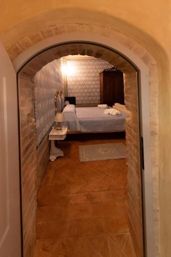 A narrow hallway leading to a bedroom with a bed, a nightstand, a lamp, and a wooden wardrobe. - PALAZZO DEL CARDINALE (Montalcino)