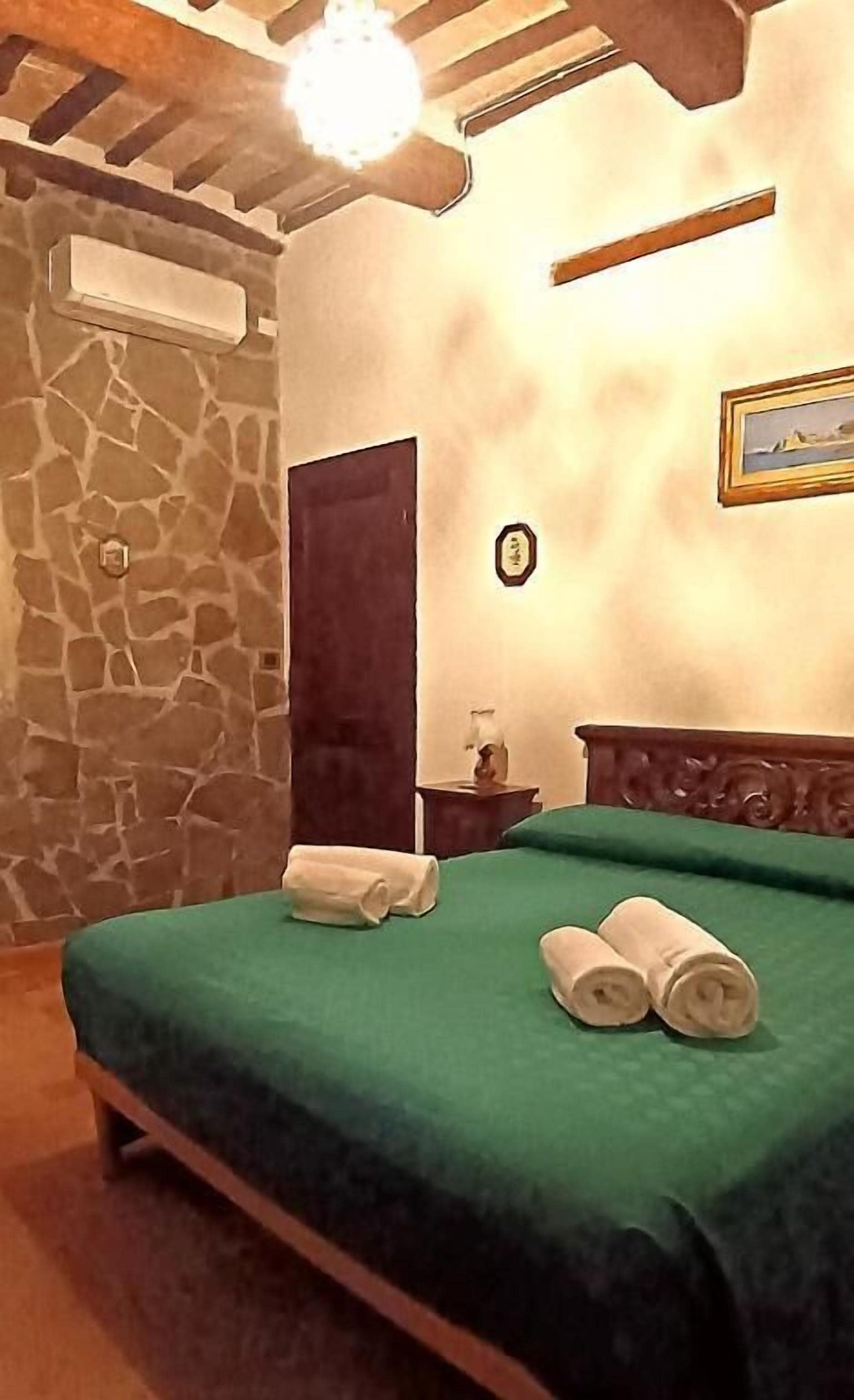 Quarto com parede de pedra, cama com roupa de cama verde, cabeceira de madeira, mesa de cabeceira e um quadro emoldurado na parede.