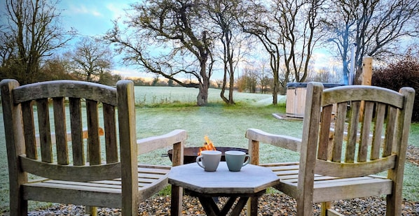 Two wooden chairs facing a table with two cups, a fire pit, and a grassy field with trees in the background.