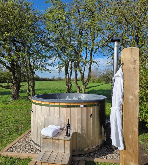 A hot tub in a wooden structure with a towel and wine bottle, surrounded by trees and a grassy field.