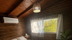 Ein Schlafzimmer einer Holzhütte mit einem Bett, einer Pendelleuchte, einem Fenster mit Vorhängen, einer Topfpflanze und einer Klimaanlage.