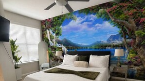 Ein Schlafzimmer mit Bett, Deckenventilator, Nachttisch, Lampe, Wandgemälde und Fenster mit Jalousien.