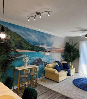 Ein Wohnzimmer mit Wandgemälde von einem Strand, einer Couch, einem Couchtisch und einem Essbereich.