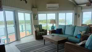 Amplia sala de estar con un gran ventanal con vistas a una masa de agua, un sofá de mimbre, un sillón a rayas y una mesa de centro.