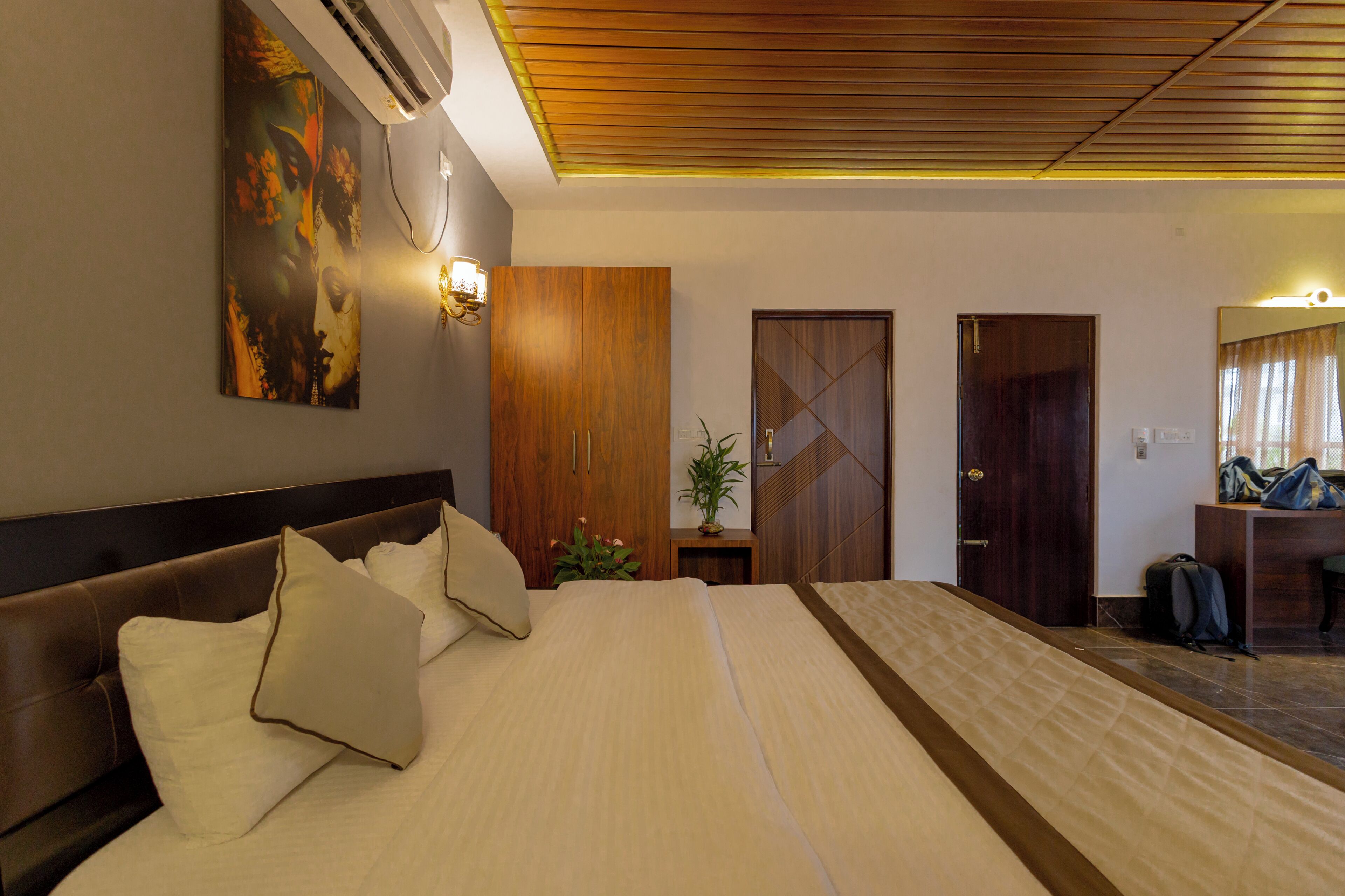 A hotel room with a large bed, wooden headboard, a wall-mounted air conditioner, a potted plant, and a mirror with a curtain.