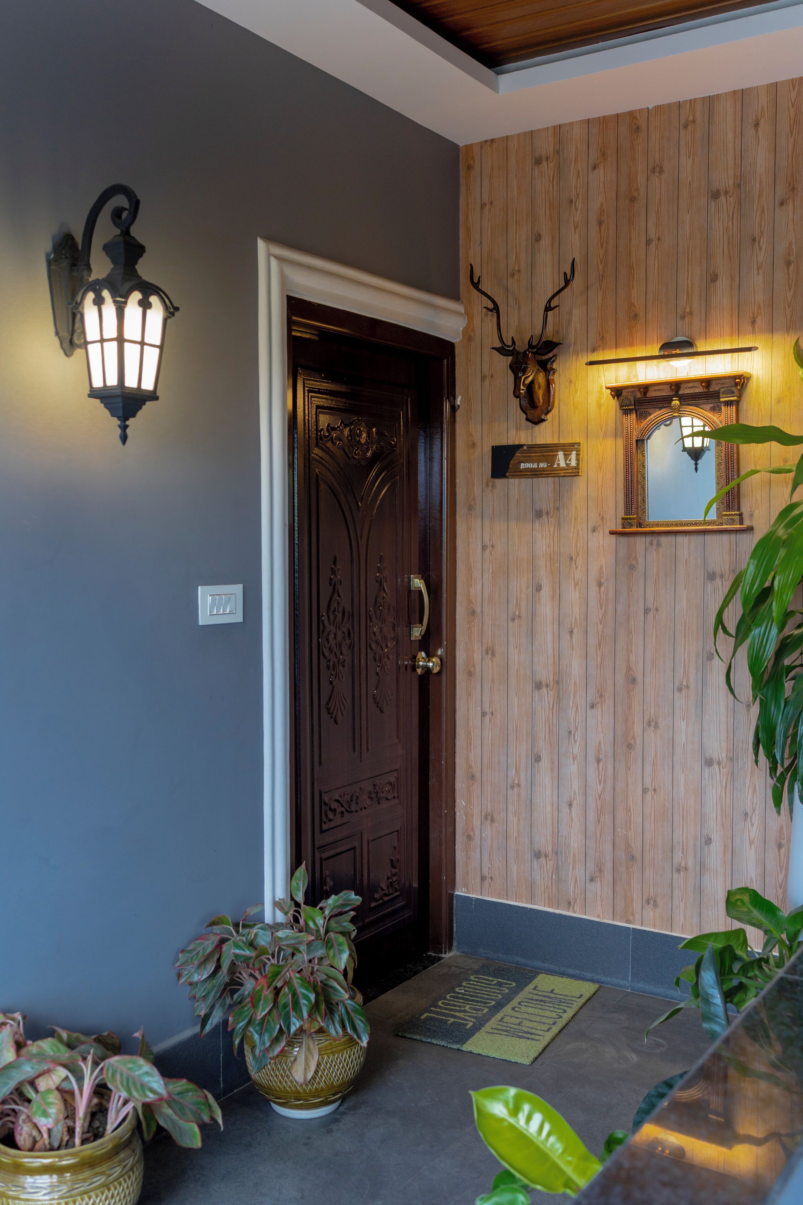 Une porte en bois avec un paillasson, un luminaire fixé au mur et une tête de cerf décorative.