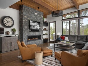 A modern living room with a stone fireplace, leather furniture, and a large window offering a view of trees. - NEW! 6 Rolling Ridge 4-Bedroom Home with Mountain view (Big Sky)