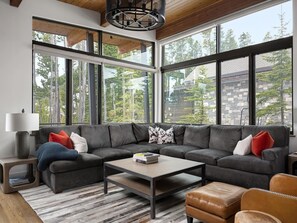 A modern living room with a sectional sofa, a coffee table, and a view of trees through large windows. - NEW! 6 Rolling Ridge 4-Bedroom Home with Mountain view (Big Sky)