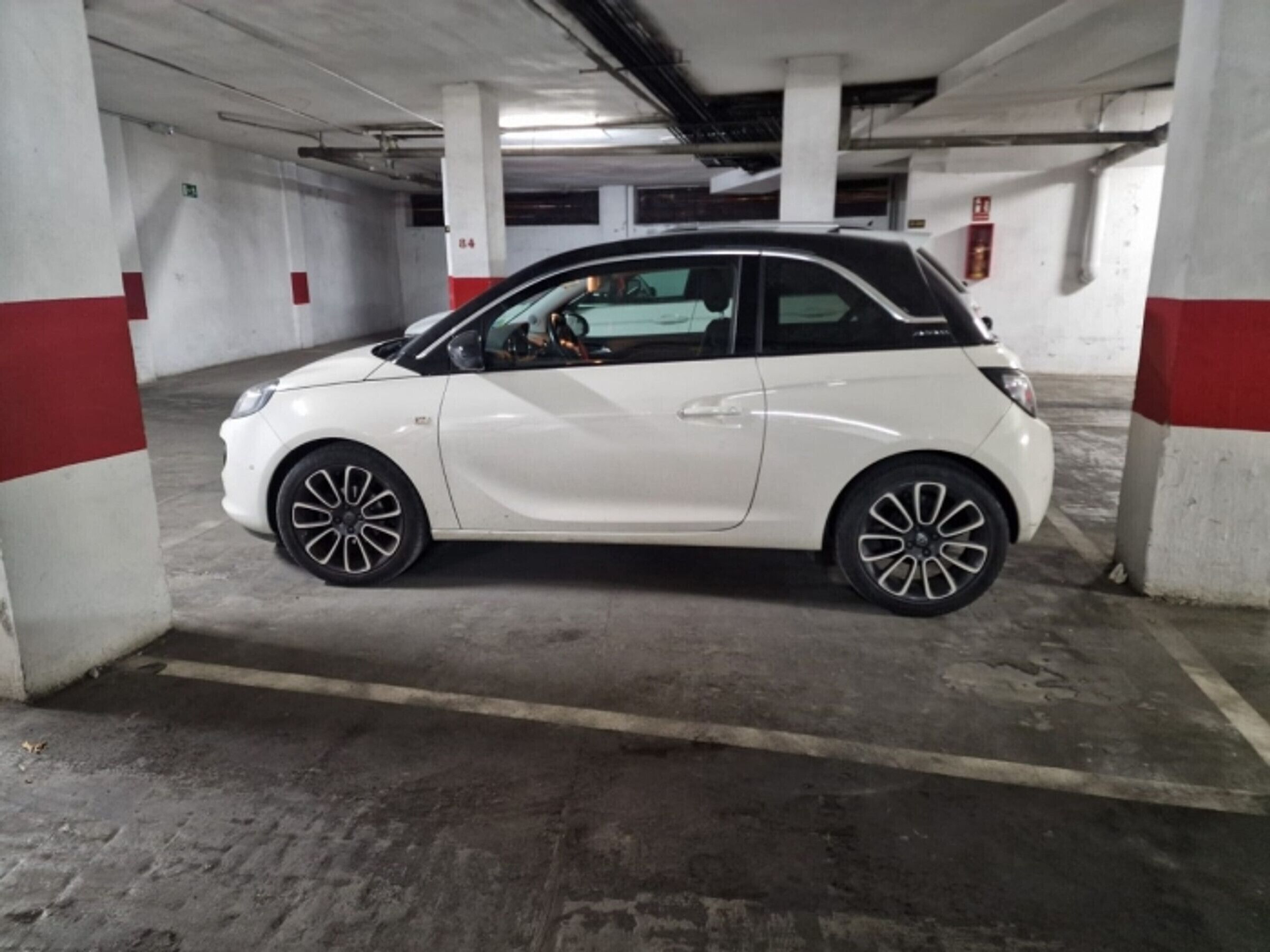 Un'auto compatta bianca parcheggiata in un parcheggio sotterraneo con pilastri in cemento e pavimento piastrellato.