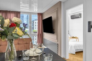 Ein Hotelzimmer mit Bett, Wandfernseher, Esstisch mit Gläsern und Blumen in einer Vase.