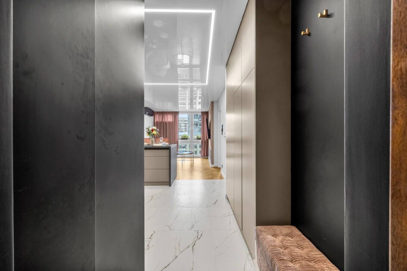 Un pasillo moderno con piso de mármol, un banco y vista hacia una cocina con mesa y sillas.