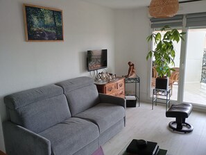 Ein Wohnzimmer mit grauem Sofa, Fernseher, einem kleinen Tisch mit Dekoration und einer Zimmerpflanze.