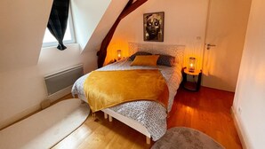 Ein Schlafzimmer mit schrägen Wänden, einem Bett mit gemusterten Textilien, einem Nachttisch mit Lampe und einem Fenster mit Vorhängen.