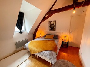 Ein Schlafzimmer mit schrägen Wänden, einem Bett mit gemusterten Textilien, einem Nachttisch mit Lampe und einem Fenster mit Vorhängen.