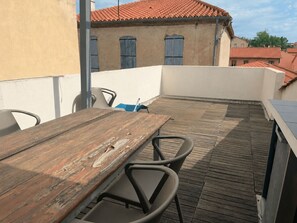 Eine Holzterrasse mit Tisch und Stühlen, ein Gebäude mit rotem Ziegeldach und eine weiße Wand.