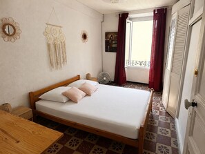 A bedroom with a wooden bed, a bedside table, a wall hanging, a window with red curtains, and a door with a handle. - Charming city center house - 8 guests (Port-Vendres)