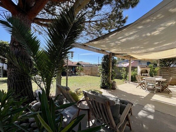 A covered patio area with a table and chairs, surrounded by plants and a tree.