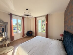 A bedroom with a large bed, a chair, a mirror, and two windows with curtains.