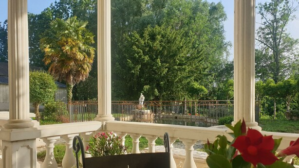 A glass table with a black sign featuring a cup and steam design, a white balustrade, and a view of a garden with trees and a statue. - Cottage by the water - 8 guests (Chérancé)