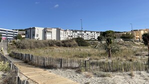 Eine Küstenlandschaft mit Holzsteg, sandigem Gelände und modernen Gebäuden im Hintergrund.