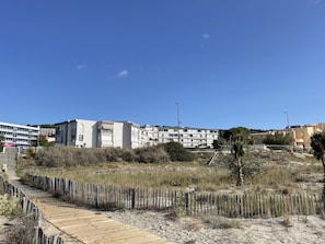 A coastal area with a wooden boardwalk, sandy terrain, and modern buildings in the background. - Studio cabin beachfront, parking - 2 guests (Sète)