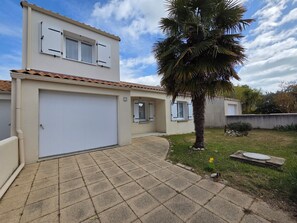 Un bâtiment à deux étages avec un garage, un petit jardin agrémenté d’un palmier et une allée pavée.