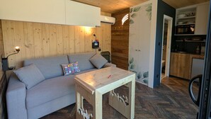 Un cuarto acogedor con techo de madera, un sofá gris con un cojín colorido, una mesita de madera y una kitchenette con armarios de madera.
