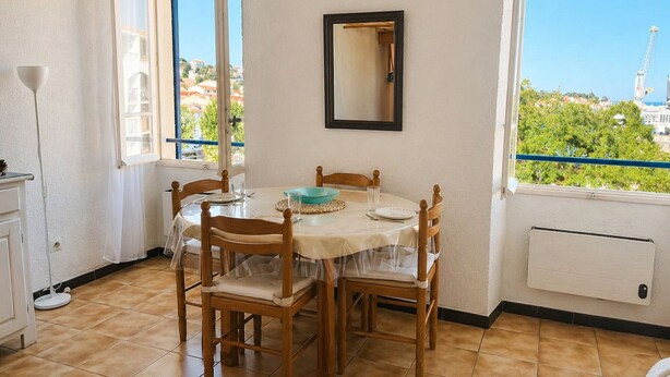 Een eetruimte met een tafel gedekt voor vier personen, houten stoelen, een raam met uitzicht op gebouwen en een kraan, en een radiator aan de muur.