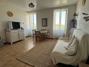 Ein Wohnzimmer mit einer Couch, einem Fernseher auf einem Schrank, einem Esstisch mit Stühlen und Blick durch die Fenster.