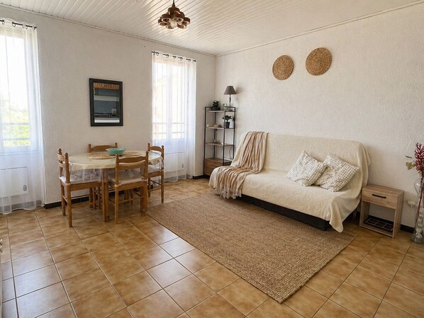 A living room with a sofa, a dining table with chairs, a lamp, and a shelf with plants. - Charming studio port view - 3 guests (Port-Vendres)
