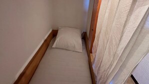 Een kleine, smalle kamer met een enkel bed, een houten hoofdbord en een dunne gordijn.