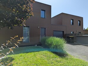 Ein modernes, zweistöckiges Gebäude mit flachem Dach, großen Fenstern und einem kleinen Vorgarten mit Rasen und Büschen.