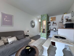 Ein Wohnzimmer mit einer Couch, einem Couchtisch, einem Esstisch und einer Küchenzeile mit einer Mikrowelle. An der Wand hängt eine Uhr.