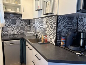 A kitchen with patterned tiles, a dishwasher, a coffee machine, and a bottle of sauce. - Apartment with sea view - 4 guests (Fleury-Saint-Pierre-la-Mer)