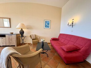 Ein Wohnzimmer mit einem roten Sofa, einem Korbstuhl, einem kleinen Tisch mit einer Lampe, einem gerahmten Bild an der Wand und einem Fliesenboden.