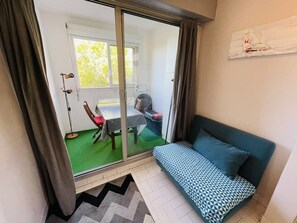 Ein Zimmer mit einer Schiebetür zum Balkon, einem Bett mit gemusterten Betttextilien, einem Sessel, einem Tisch mit Tischdecke und einem Ventilator.