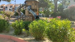 Un parque infantil con tobogán y estructuras para escalar, rodeado de vegetación y edificios residenciales.