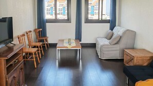 Habitación amoblada con televisión, sillas de madera, sofá, una mesita y dos ventanas con cortinas.