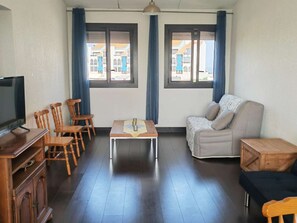 A furnished room with a television, wooden chairs, a sofa, a small table, and two windows with curtains. - Air-conditioned apartment - relaxing setting (Le Barcarès)