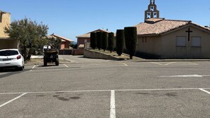 Un parking avec une voiture blanche et un quad, un bâtiment surmonté d’une croix, et un ciel bleu dégagé.