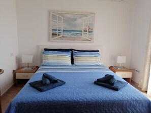 A bedroom with a bed, two bedside tables, a painting of a beach scene, and a view of the sea. - MOYS APARTMENT SEA VIEW OLBIA (Olbia)