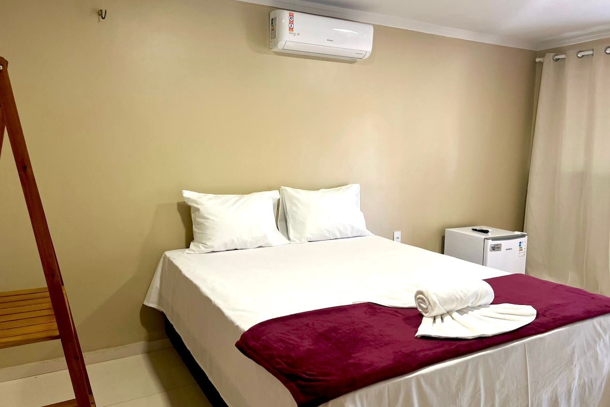 A hotel room with a bed, a white air conditioner, a small bedside table, and a curtain.