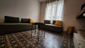 Una habitación con piso estampado, un sofá con cojines, una mesita y una ventana con cortinas.