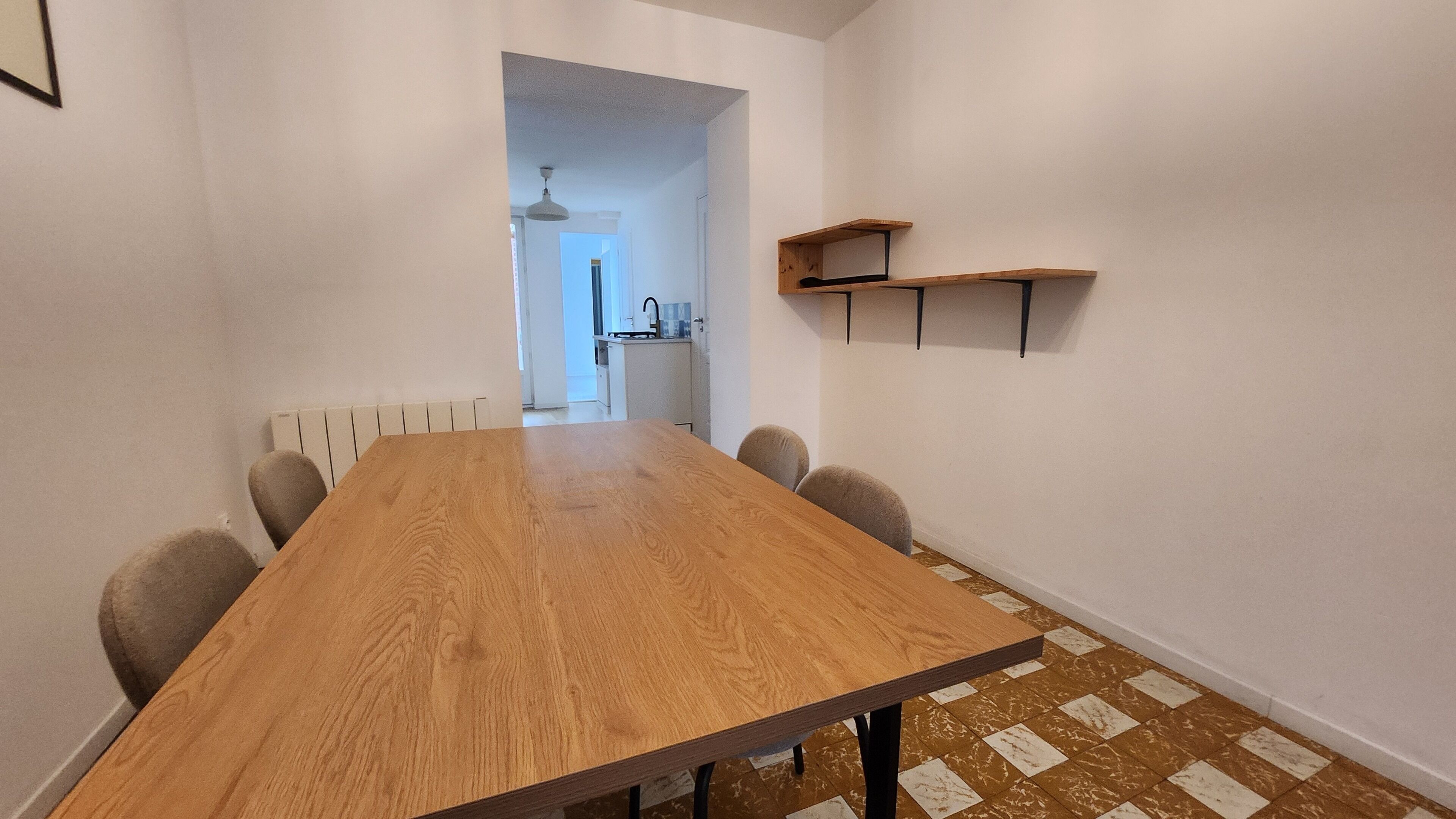 Une longue table en bois, entourée de chaises, un radiateur, une étagère avec divers objets, et un coin cuisine à l’arrière-plan.